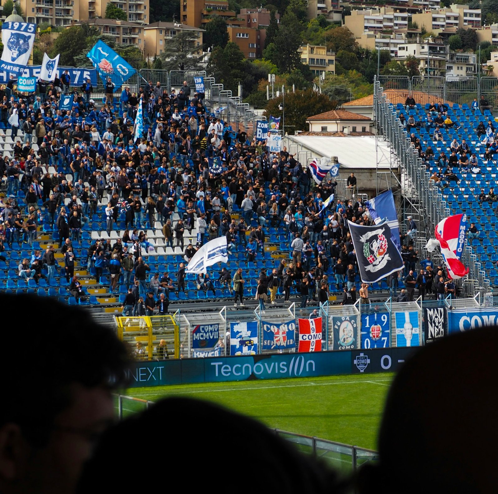 Como 1907 supporters standing among blue seats and flags at Stadio Giuseppe Sinigaglia